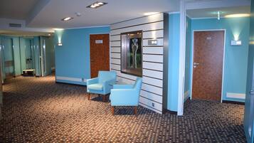 Un vestíbulo de hotel con alfombra estampada, paredes azul claro, un mostrador de recepción, dos sillones y un cuadro en la pared.