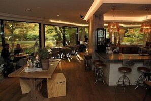 A modern restaurant with wooden tables and chairs, large windows with a view of trees, and a bar area with stools. - Congress Hotel Am Stadtpark (Hannover)
