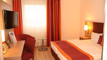 Ein Hotelzimmer mit einem Bett, einem Schreibtisch mit Stuhl, einem Fernseher und einem Fenster mit Vorhängen.