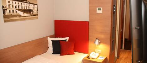 A hotel room with a bed, a nightstand with a lamp, a telephone, and a door leading to a hallway.