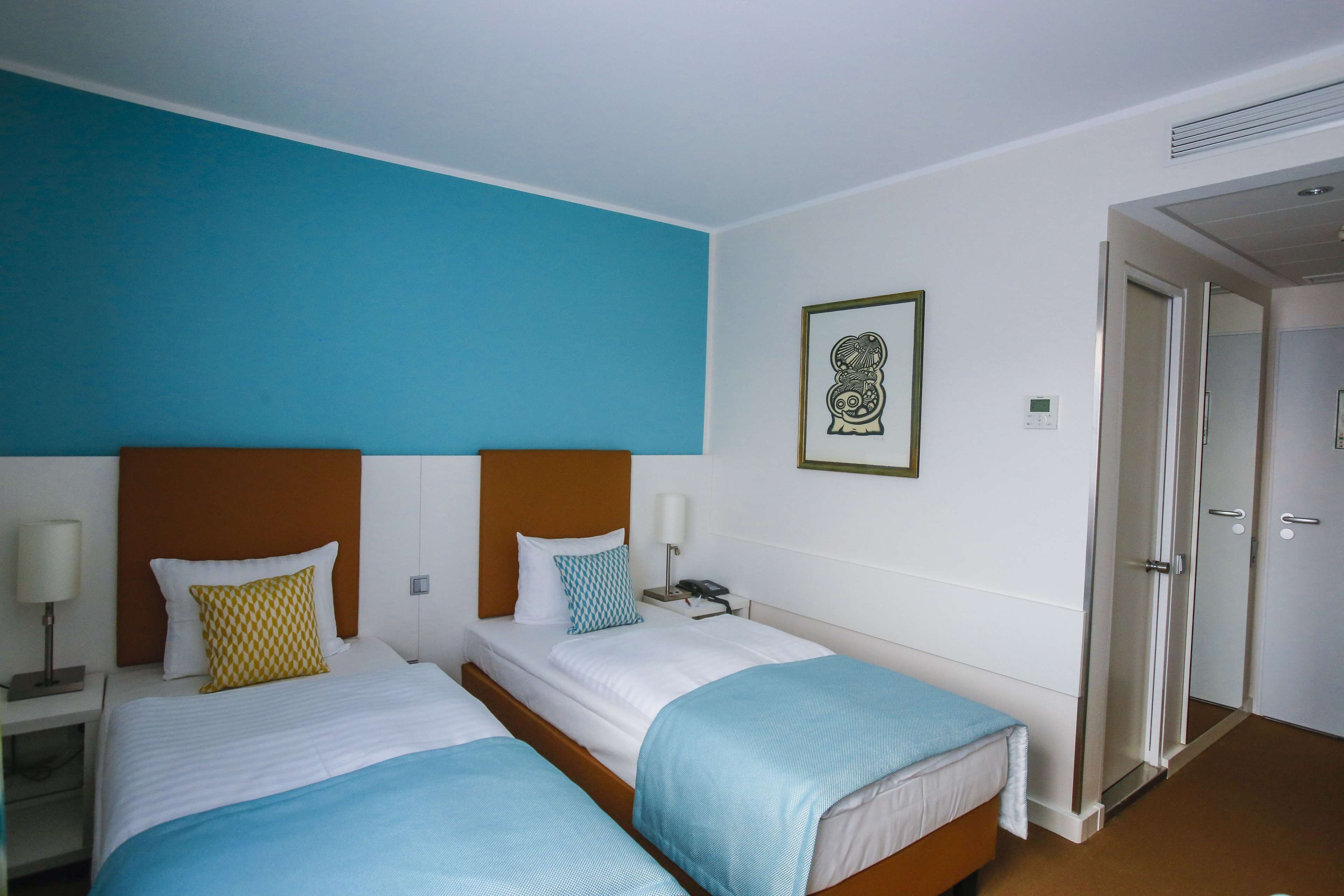 Een hotelkamer met twee bedden, een nachtkastje met een lamp, een lijst met een afbeelding aan de muur en een deur naar een andere kamer.
