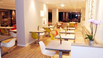 A modern restaurant with multiple dining tables and chairs, a wooden floor, and a view of the dining area through a glass wall.