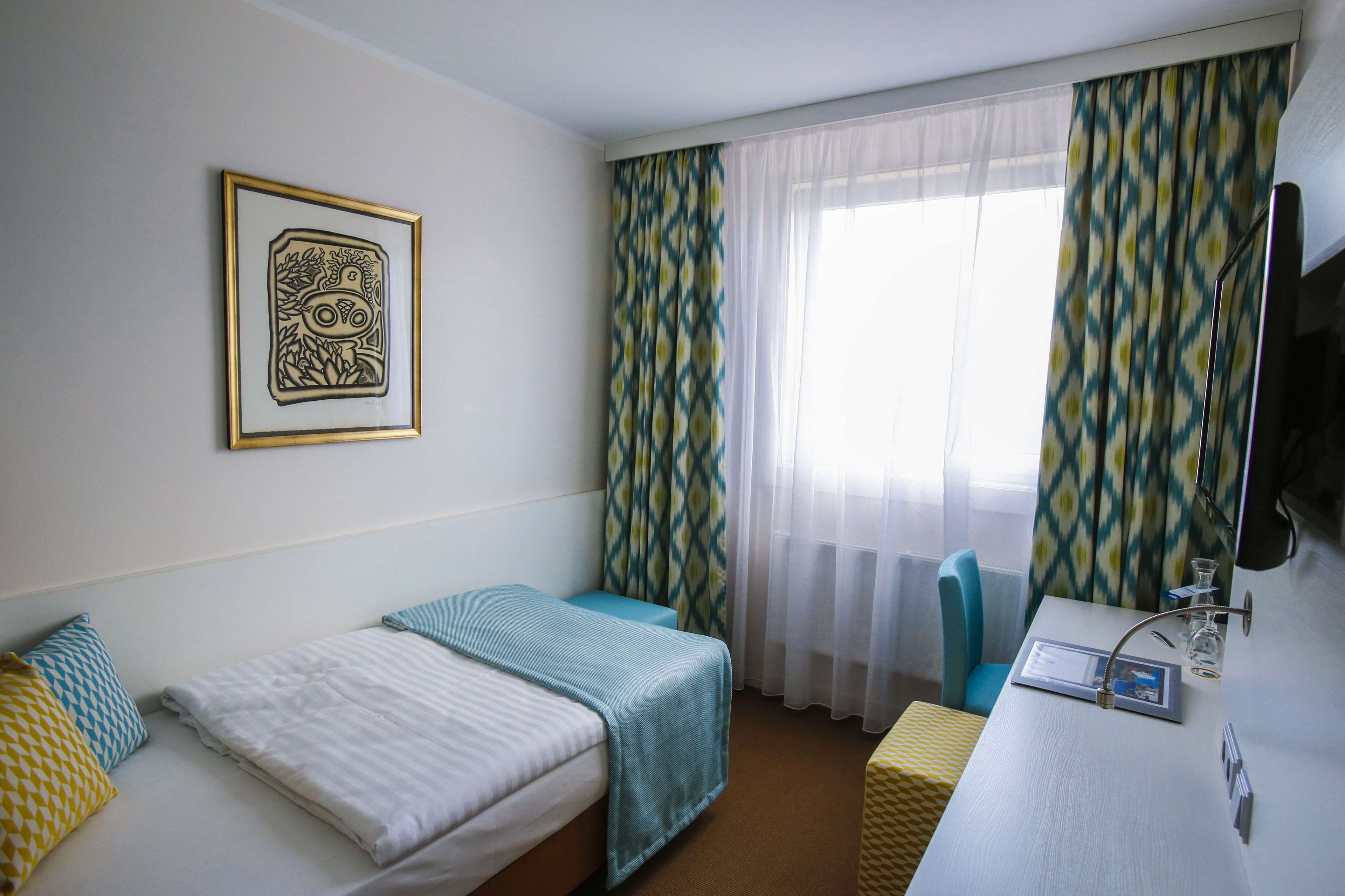 Habitación de hotel con cama, escritorio con computadora, silla y ventana con cortinas estampadas.