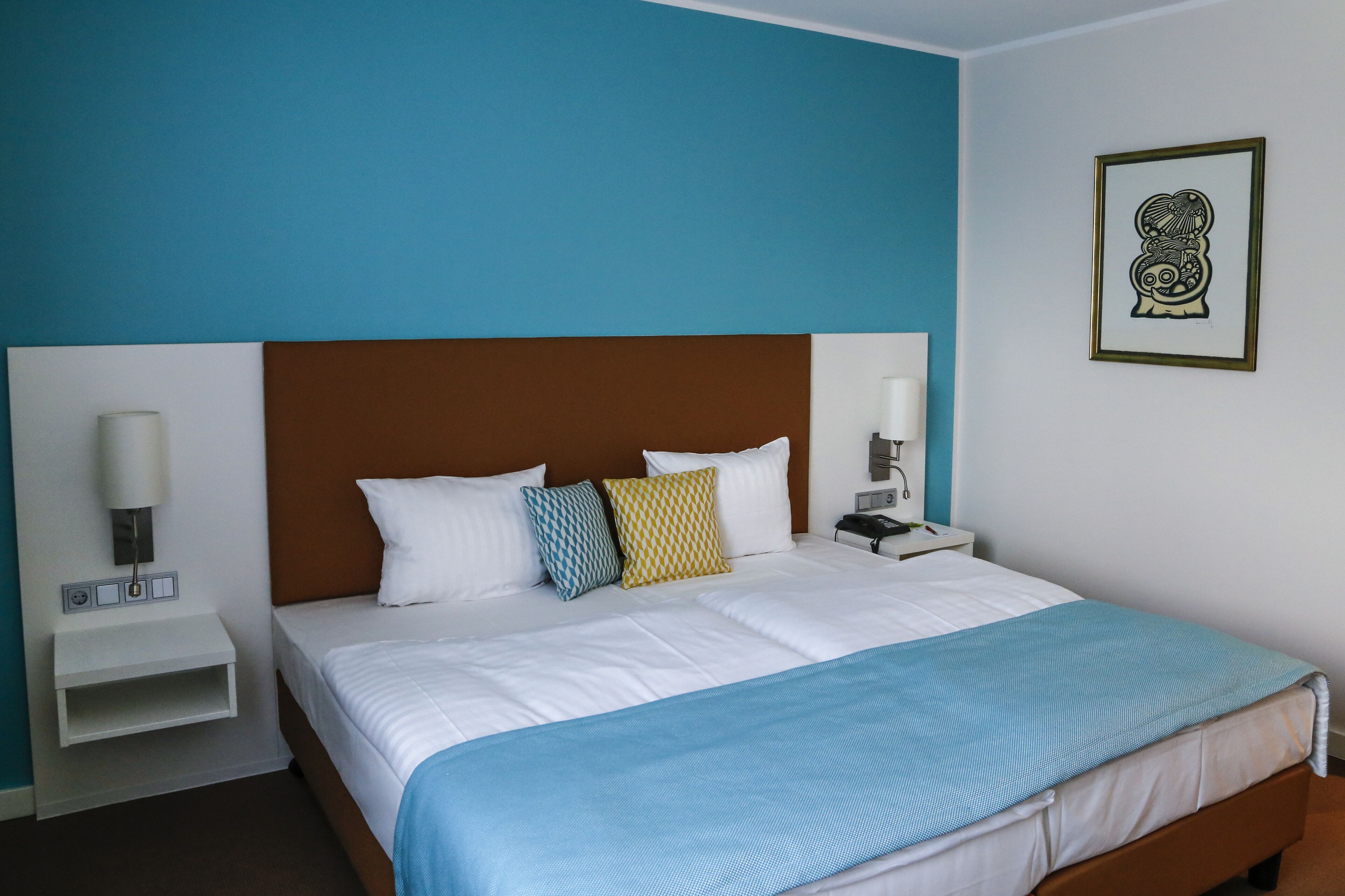 A hotel room with a large bed, white bedding, a blue and white striped blanket, and a brown headboard. There are two bedside tables with lamps and a small shelf.