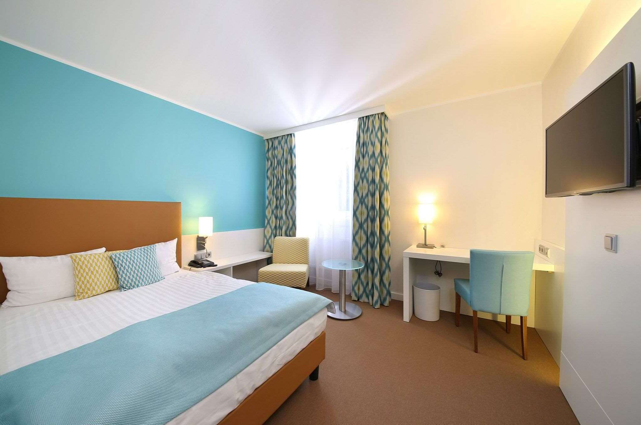 A hotel room with a bed, a desk, a chair, a TV, and a window with curtains.
