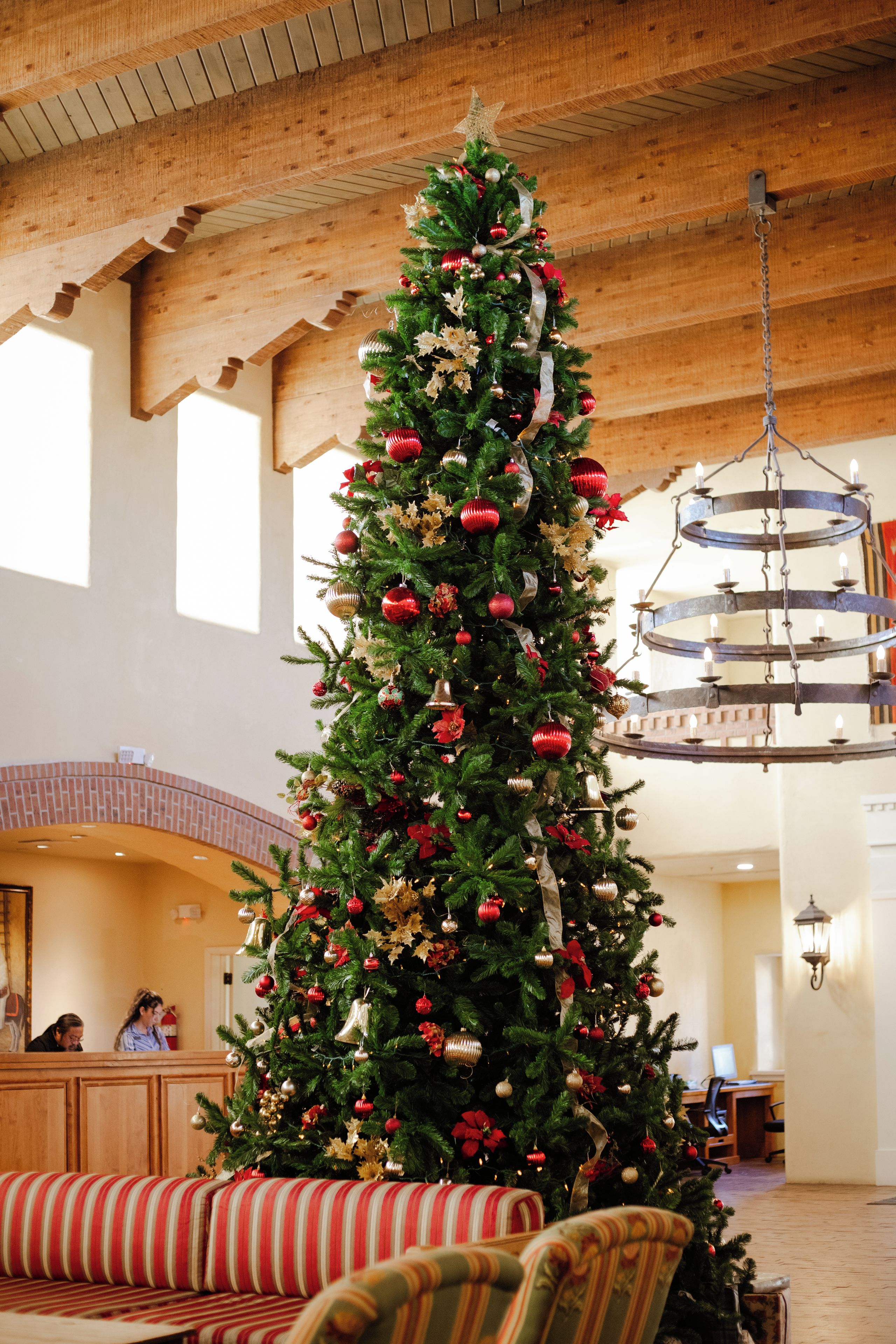 A large Christmas tree with ornaments and lights in a spacious lobby with wooden beams and a chandelier.