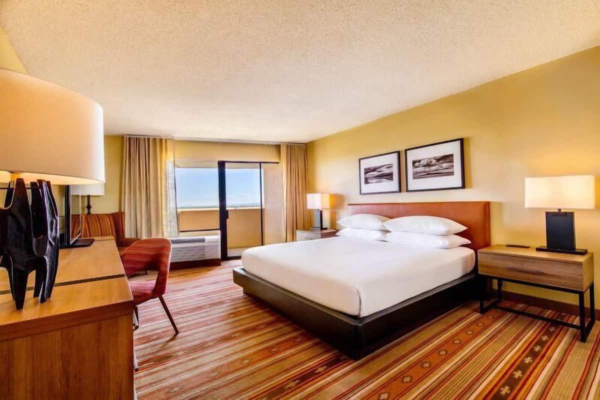 A hotel room with a bed, bedside tables, a desk, a chair, and a view of the outside.