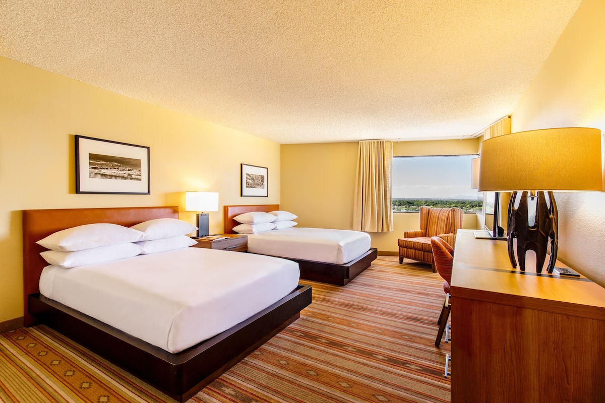 A hotel room with two beds, a desk, a chair, and a view of the outdoors.