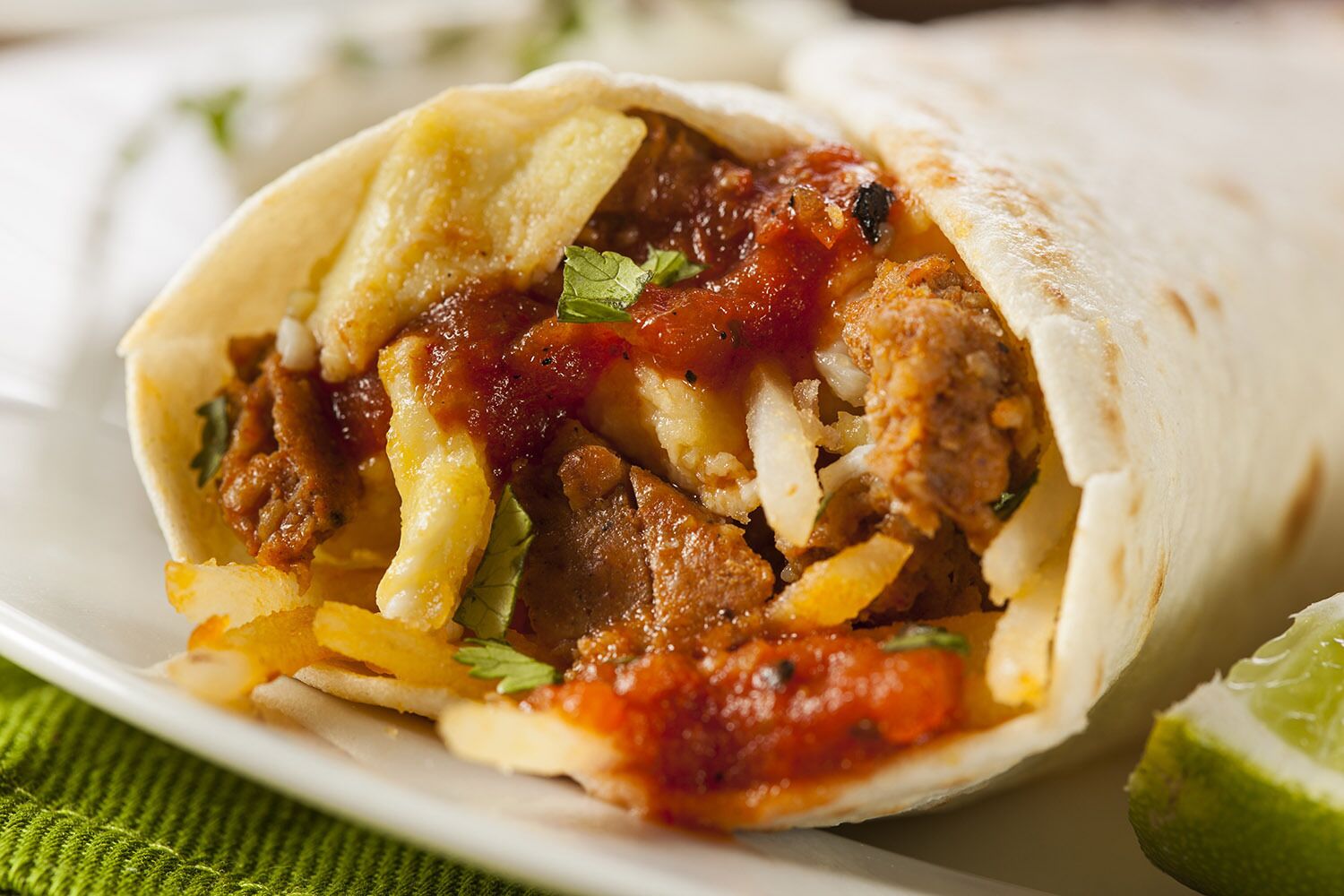 A burrito filled with meat, potatoes, and sauce, served on a white plate with a lime wedge.