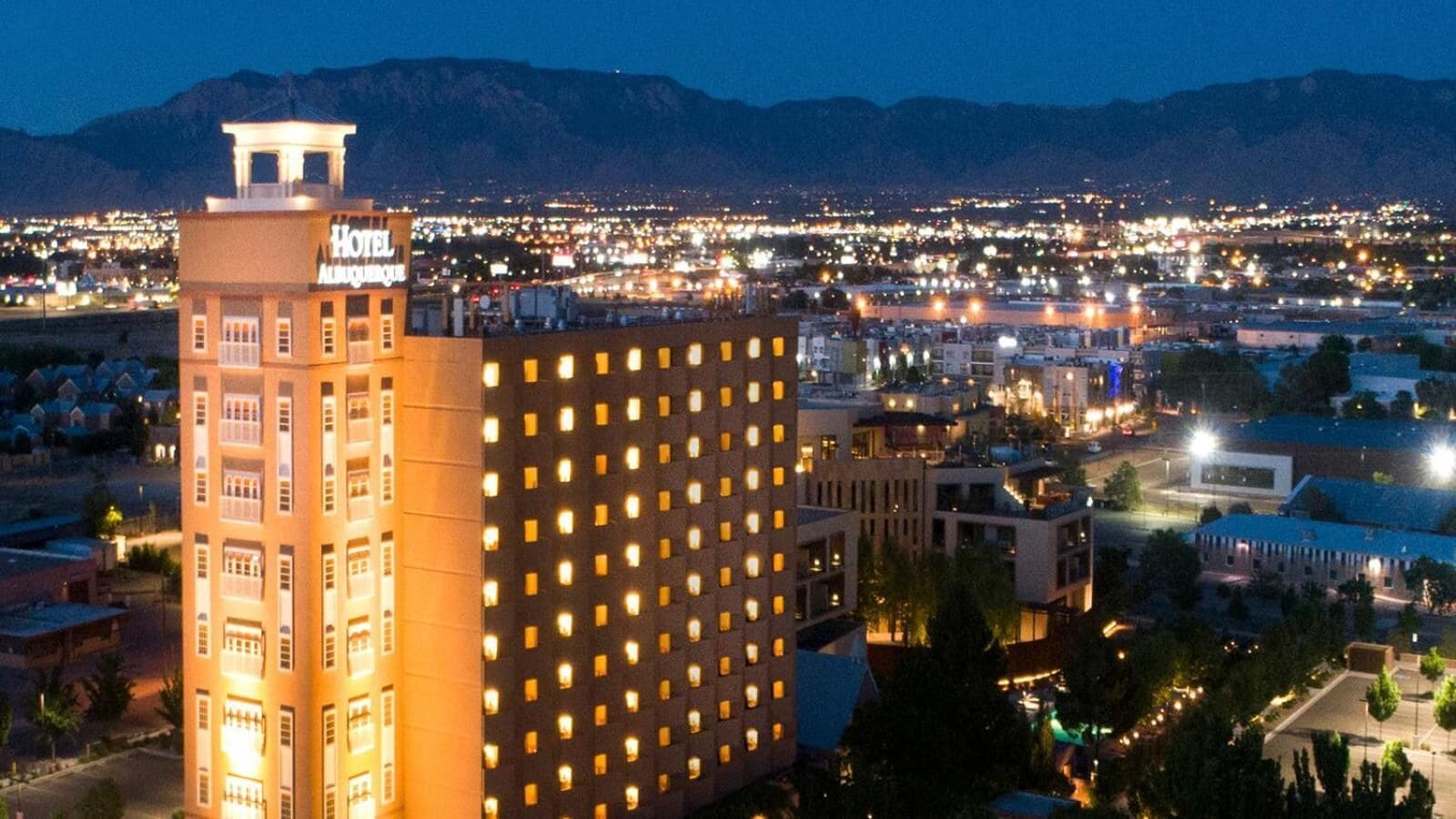 Un hotel con varios pisos y una torre destacada, rodeado por un paisaje urbano con edificios visibles y montañas al fondo.