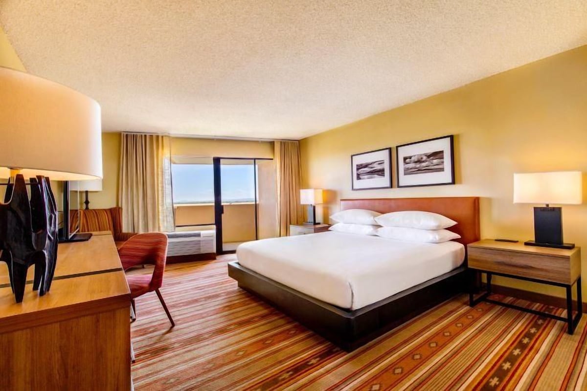 A hotel room with a bed, bedside tables, a desk, a chair, and a view of the outside.