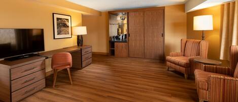 A hotel room with a wooden floor, a television, a desk with drawers, a chair, a sofa, a small table, and a lamp.