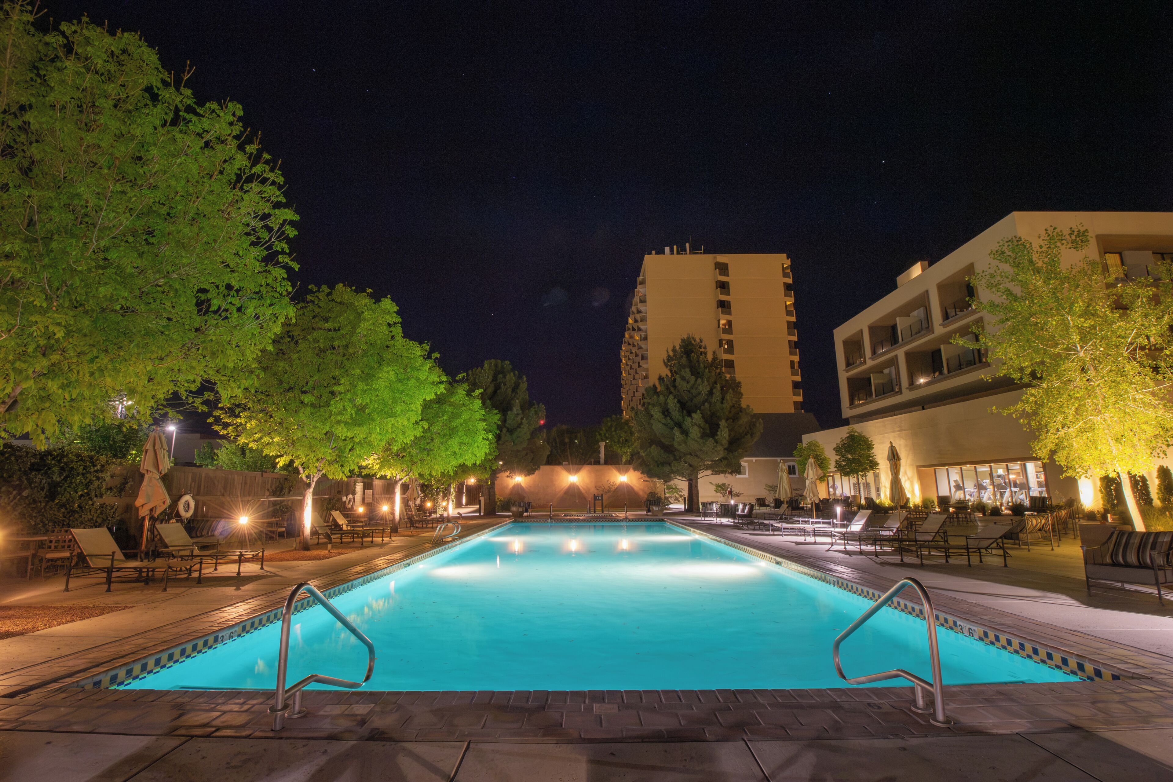 A swimming pool with a clear blue water surface, surrounded by lounge chairs and umbrellas, with trees and a building in the background.