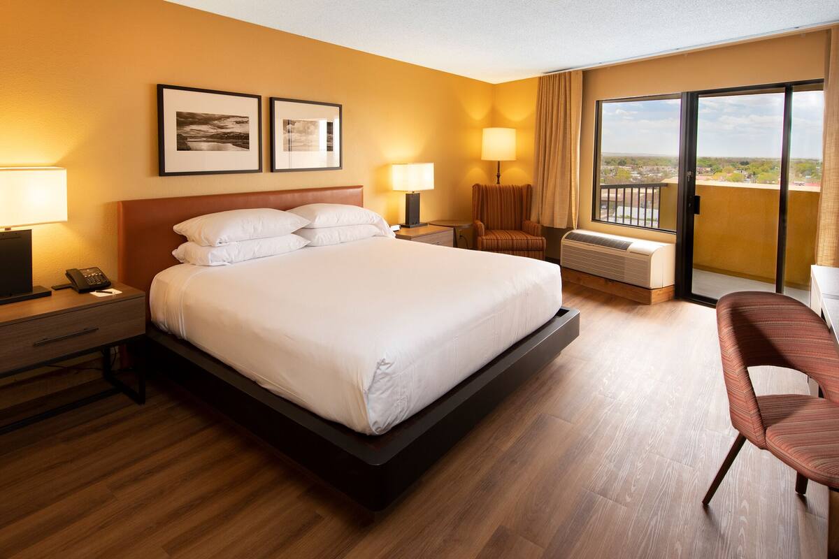 A hotel room with a large bed, a desk with a lamp, a chair, a window with a view, and a sliding door to a balcony.