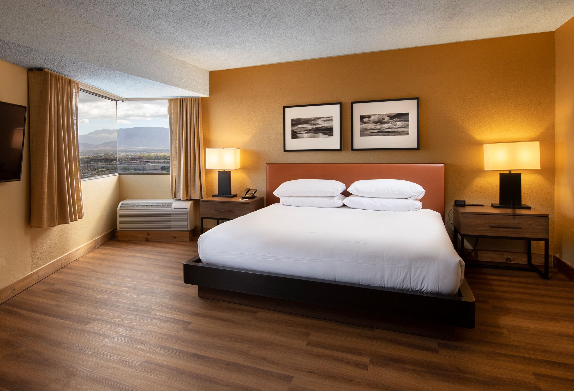 A hotel room with a large bed, two bedside tables, a flat-screen TV, a window with curtains, and two framed pictures on the wall.