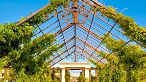 Espaço para eventos ao ar livre com estrutura de cobertura de vidro geométrica, rodeado por vegetação e mesas e cadeiras brancas.
