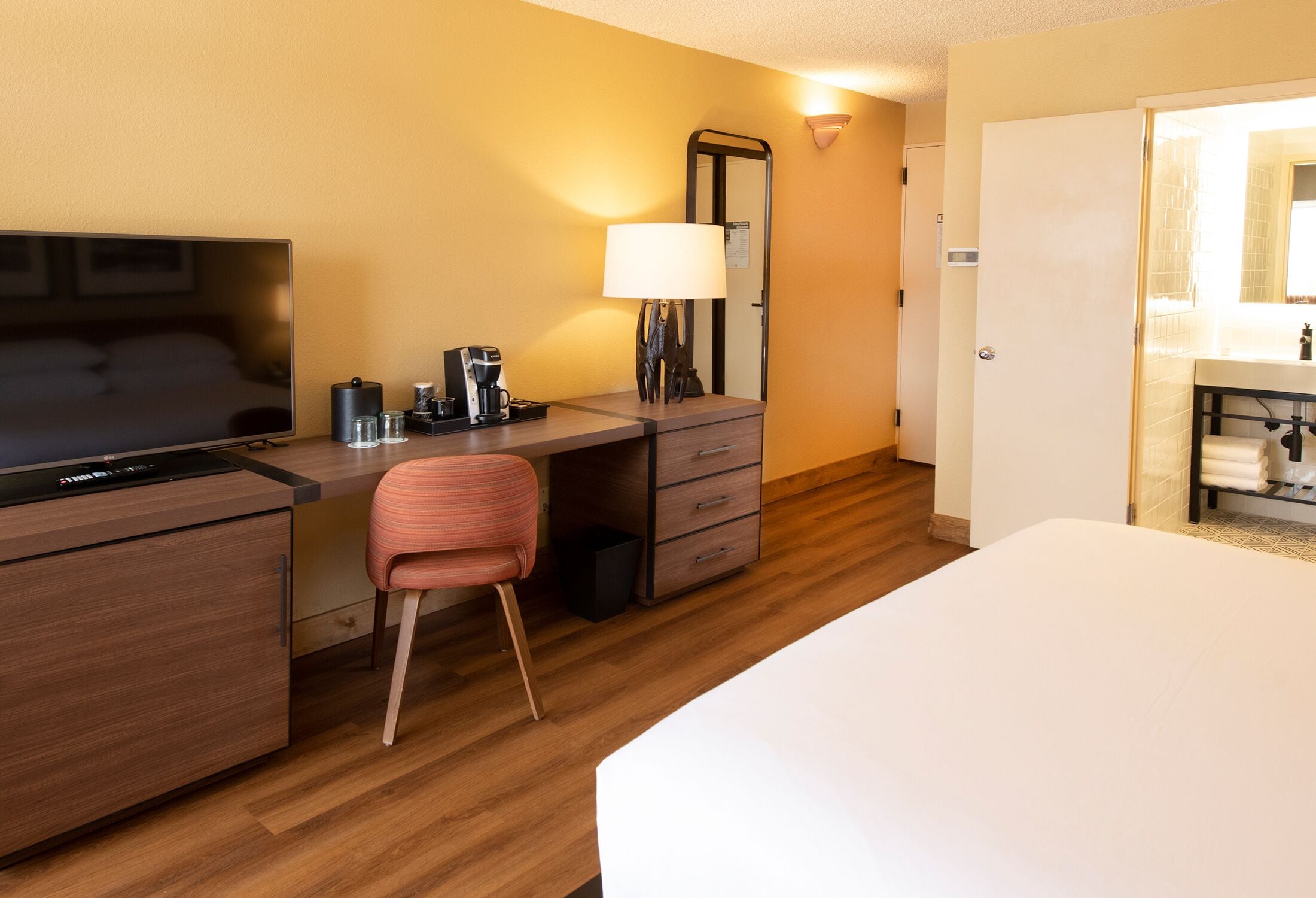 A hotel room with a bed, a desk with a chair, a TV, a lamp, a coffee maker, and a mirror.