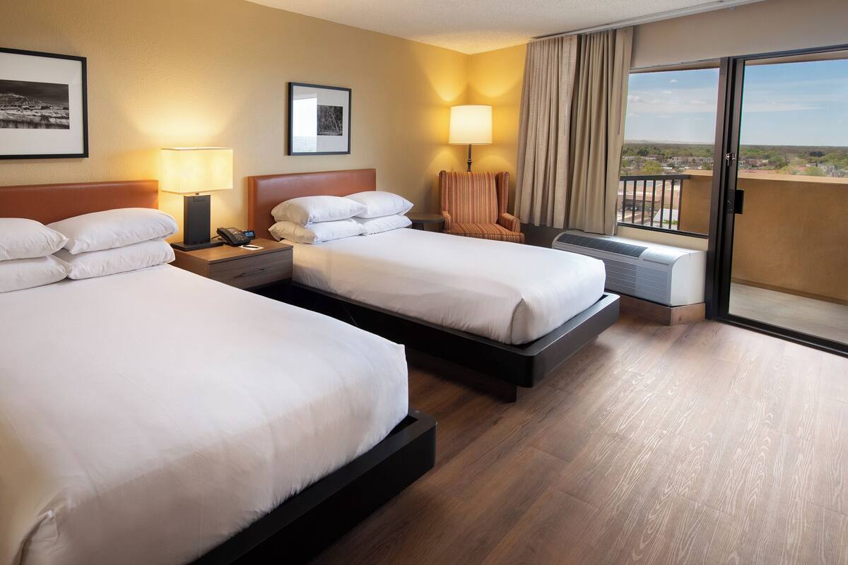 A hotel room with two beds, a chair, a desk with a lamp, and a large window with a view.