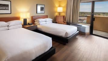 A hotel room with two beds, a chair, a desk with a lamp, and a large window with a view.