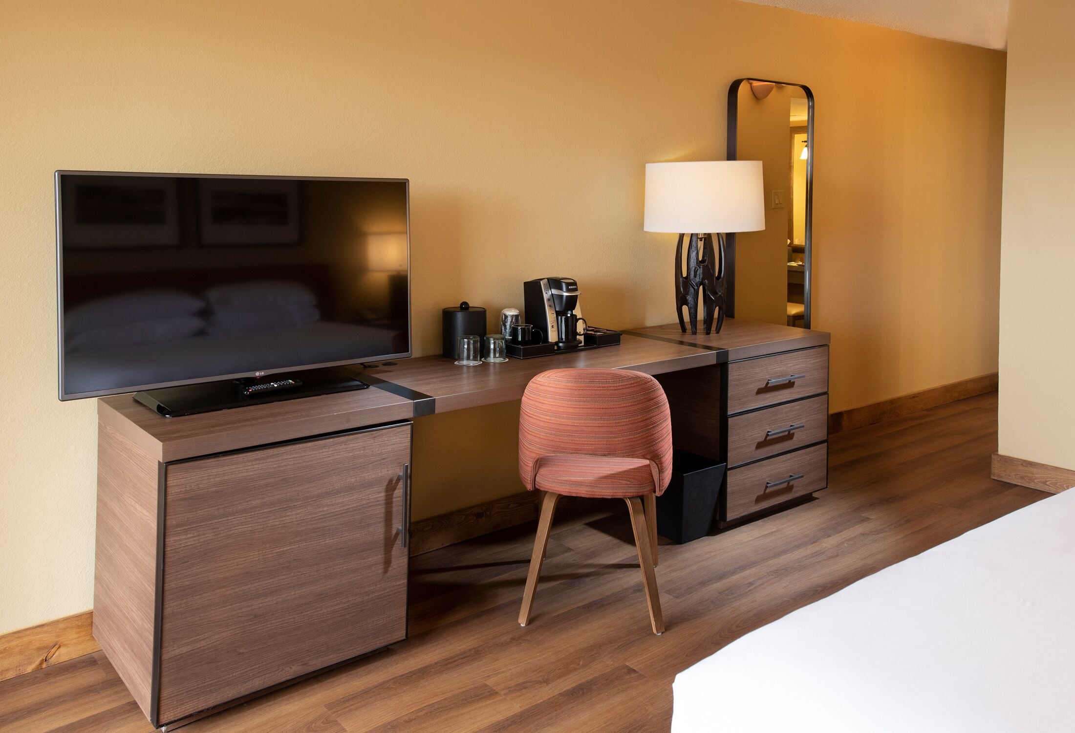 A hotel room with a TV, a desk with a coffee maker, a chair, a lamp, and a mirror.