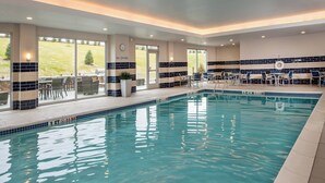 Piscina interior con agua cristalina, rodeada de sillones de descanso y mesas, y grandes ventanales con vistas al exterior.