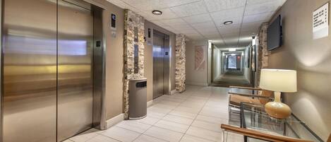A hotel corridor with elevators, a trash bin, a glass table with a lamp, and a wall-mounted TV.