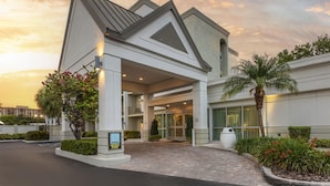 A modern hotel entrance with a covered driveway, a palm tree, and a sign indicating a parking area.