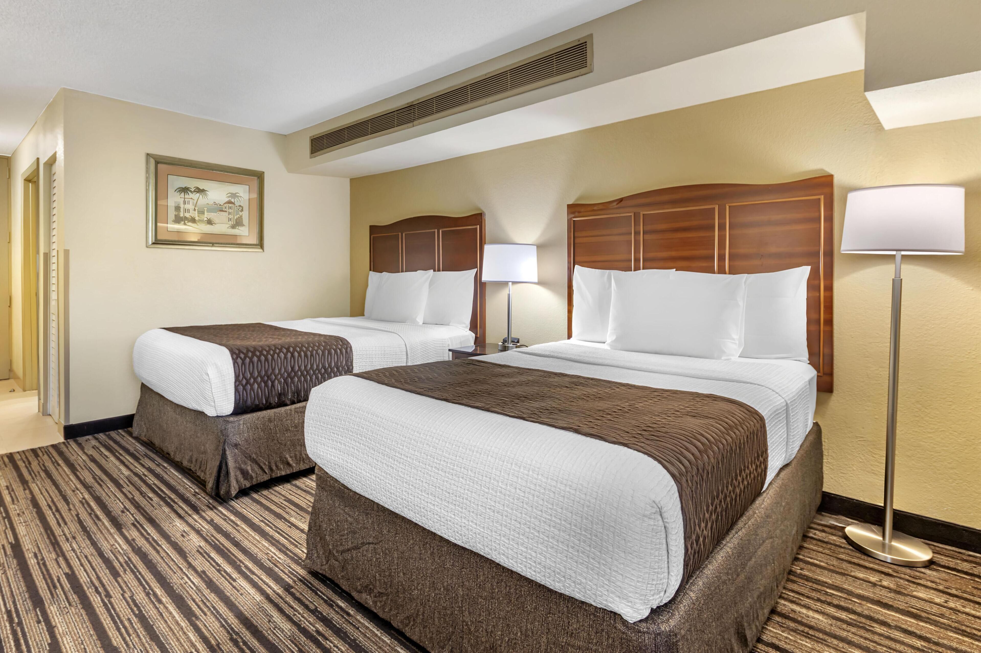 A hotel room with two beds, a large window, a framed picture on the wall, and a standing lamp.