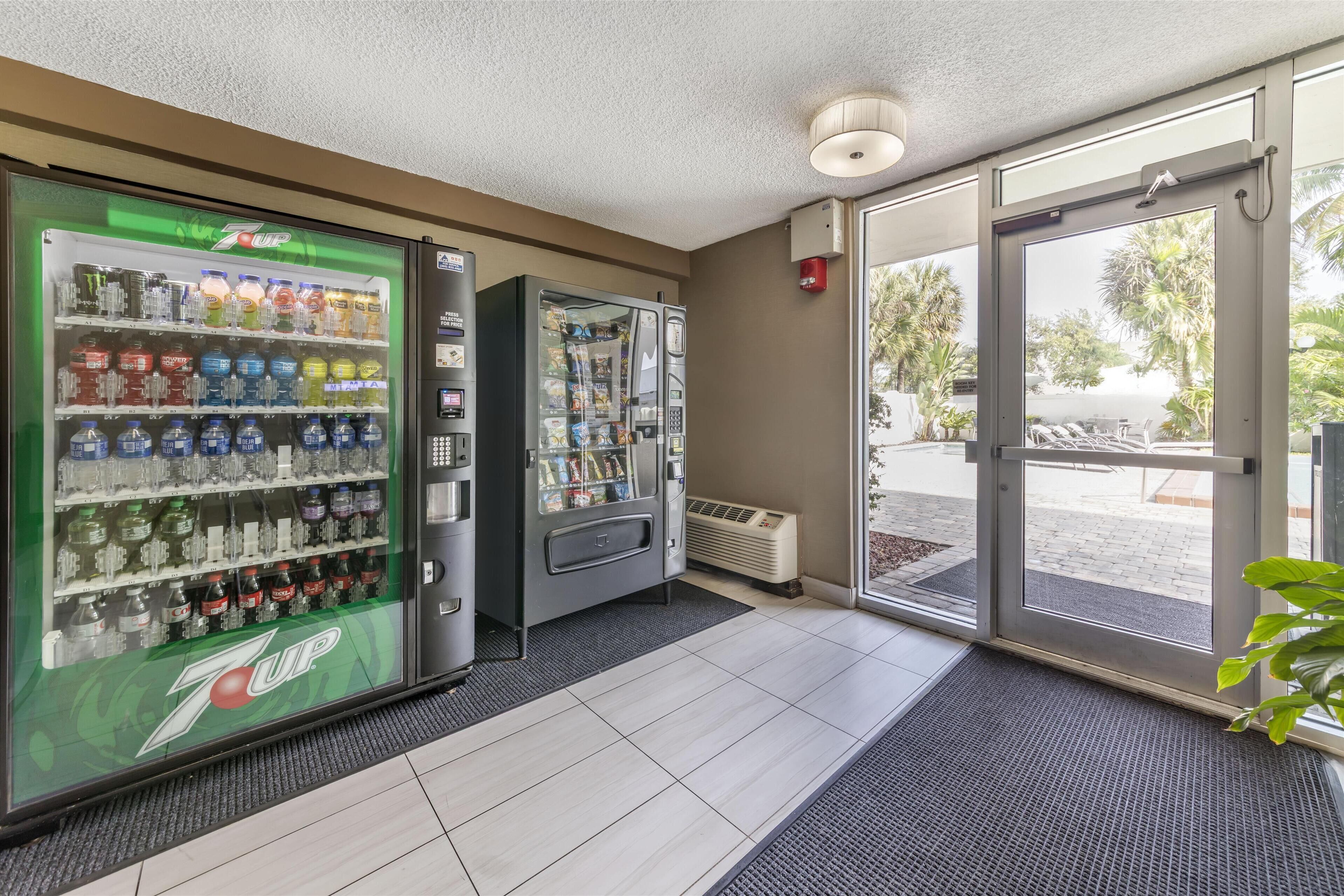 Una máquina expendedora de 7UP y un dispensador automático de bebidas en el vestíbulo de un hotel, con suelo de baldosas y vistas al exterior.