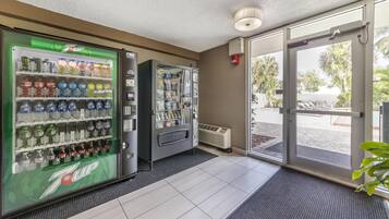 Una máquina expendedora de 7UP y un dispensador automático de bebidas en el vestíbulo de un hotel, con suelo de baldosas y vistas al exterior.