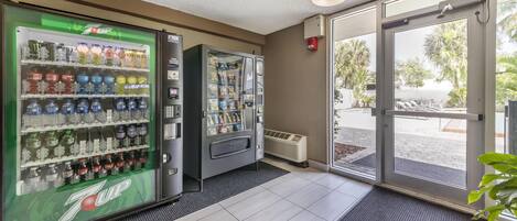 Eine 7UP-Getränkeautomaten und ein Selbstbedienungsgetränkespender in der Hotelhalle mit Fliesenboden und Blick ins Freie.