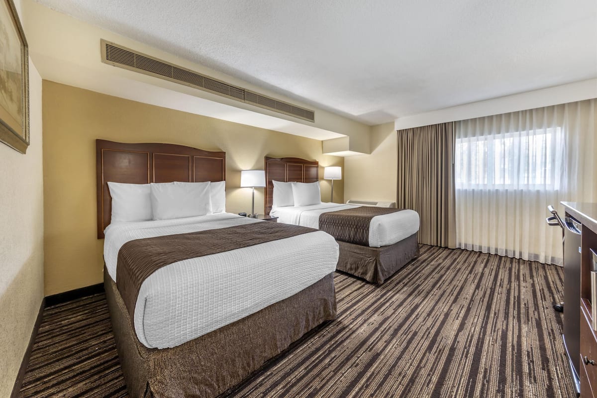 A hotel room with two beds, a large window with curtains, a carpeted floor, and a ceiling vent.