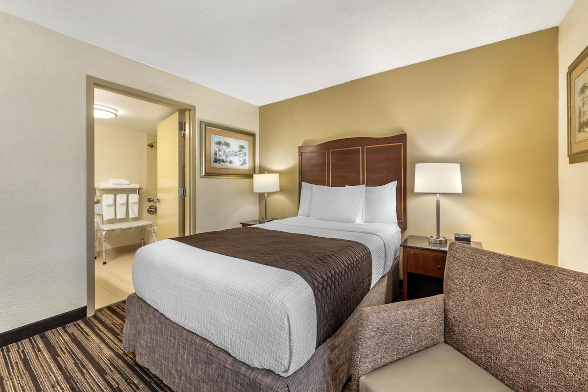 A hotel room with a bed, a nightstand, a lamp, a chair, and a bathroom visible through an open door.