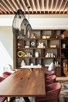 Un comedor moderno con una mesa de madera y sillas tapizadas en rojo, una estantería de madera oscura con diversos objetos decorativos e iluminación colgante.