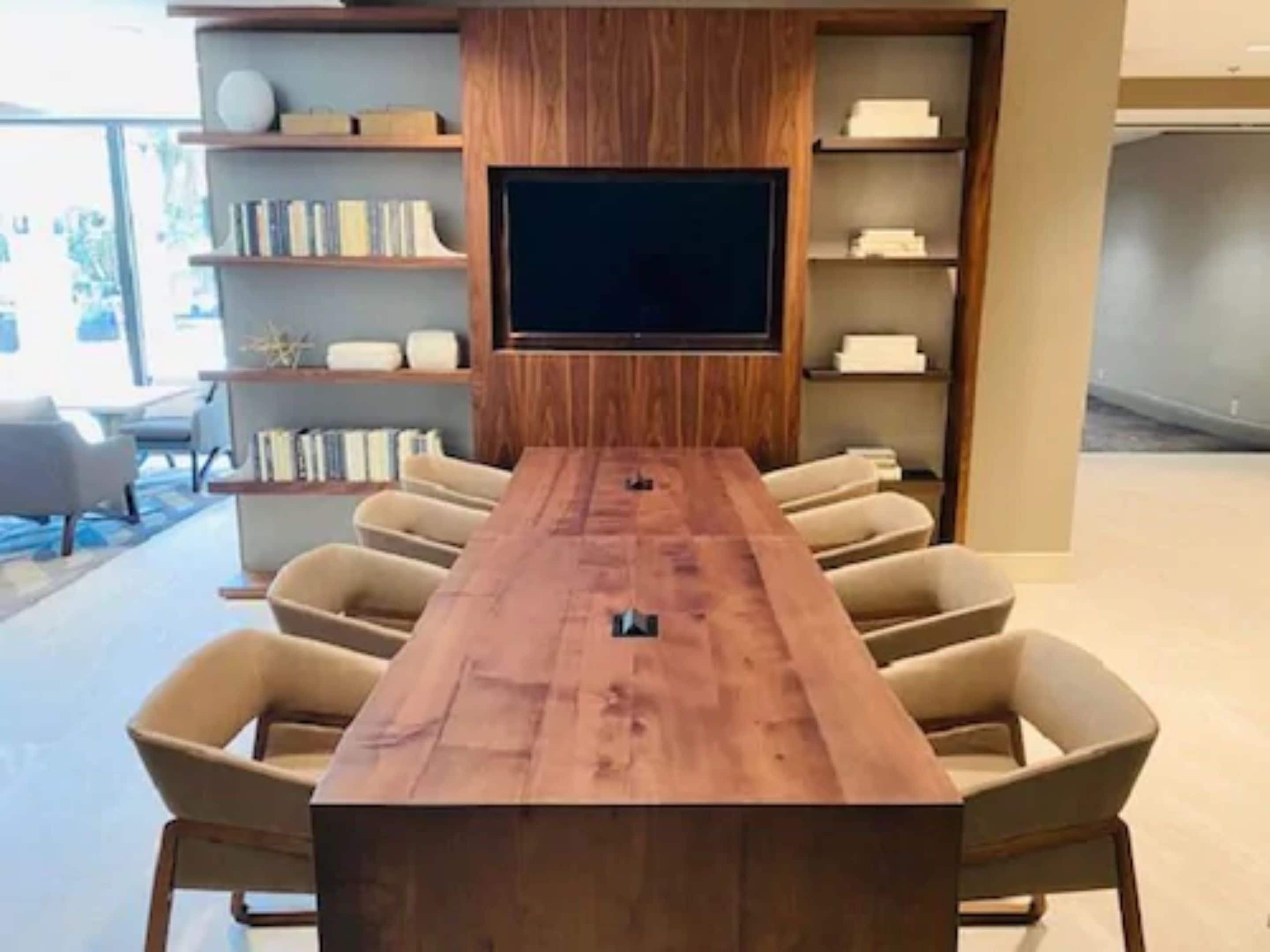 Un salón de conferencias con una larga mesa de madera, sillas color beige, un televisor de pantalla plana grande y estanterías empotradas con libros y objetos decorativos.