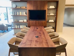 A conference room with a long wooden table, beige chairs, a large flat-screen TV, and built-in shelving with books and decorative items.