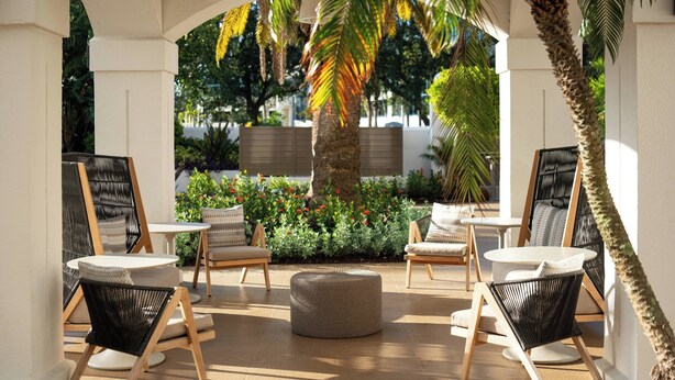 An outdoor seating area with wicker chairs and a round ottoman, surrounded by lush greenery and palm trees.