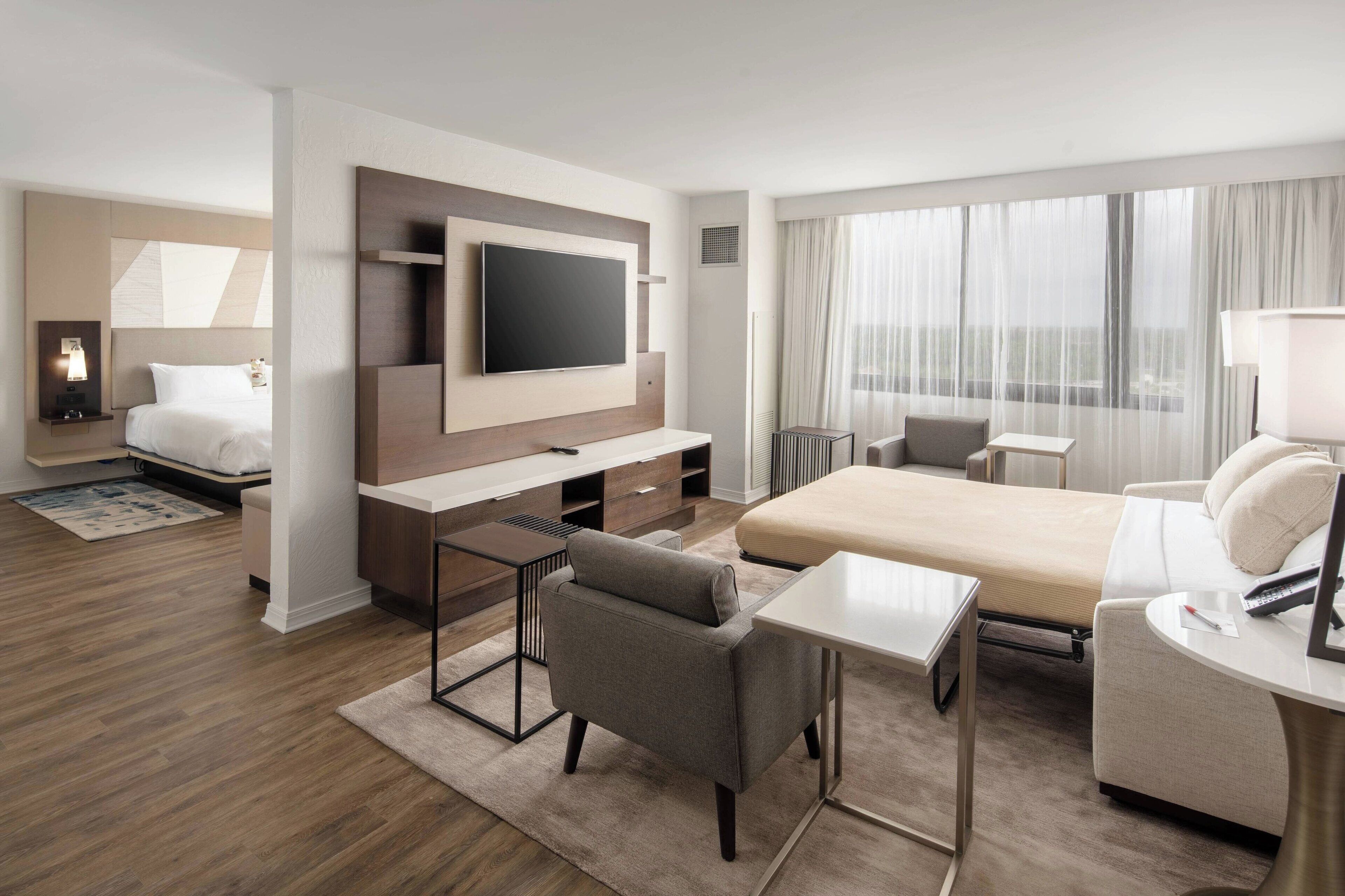 A modern hotel room with a bed, a desk, a television, and a seating area.