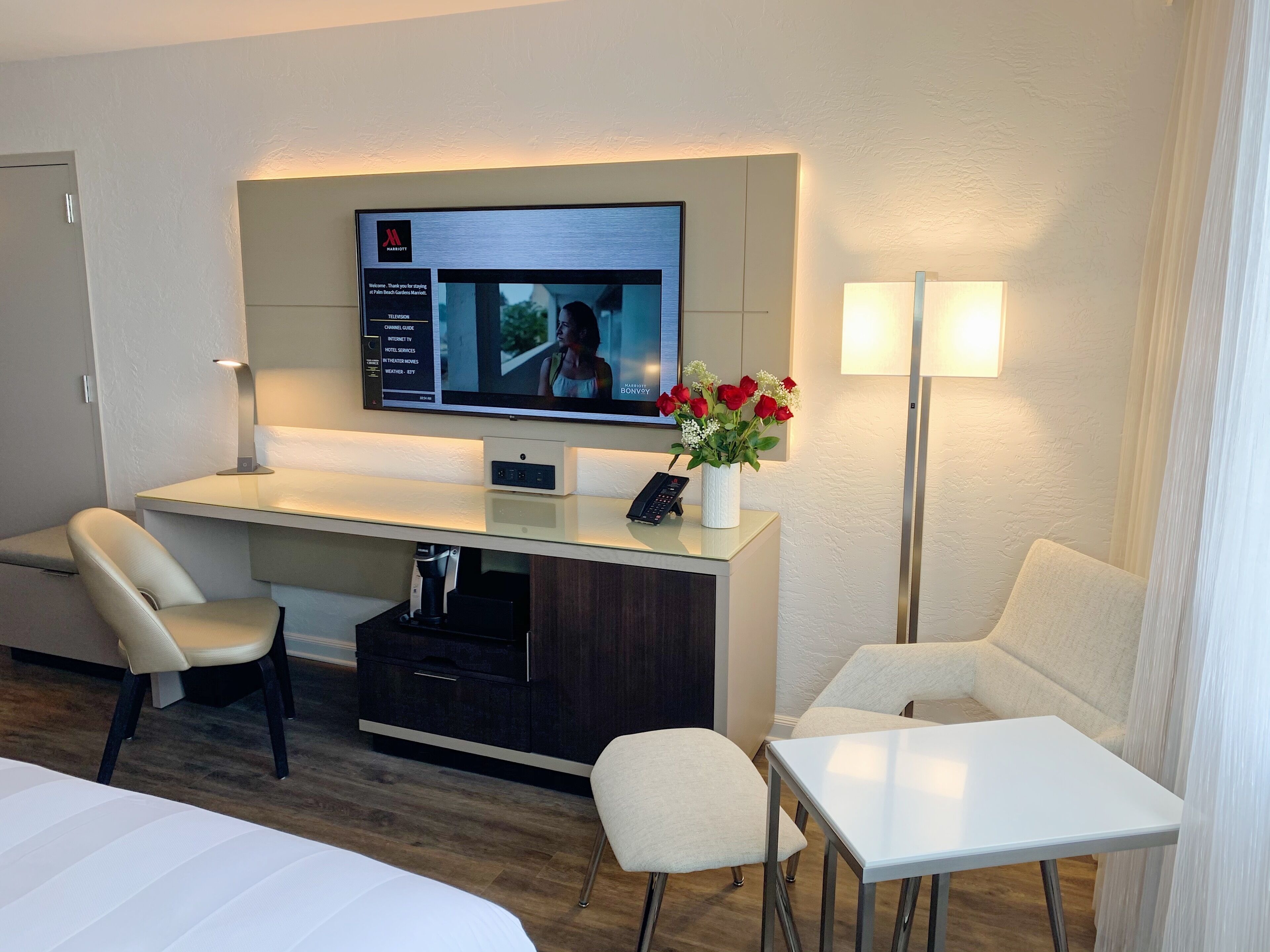 A hotel room with a flat-screen TV mounted on the wall, a desk with a vase of flowers, a chair, a bedside table, and a lamp.