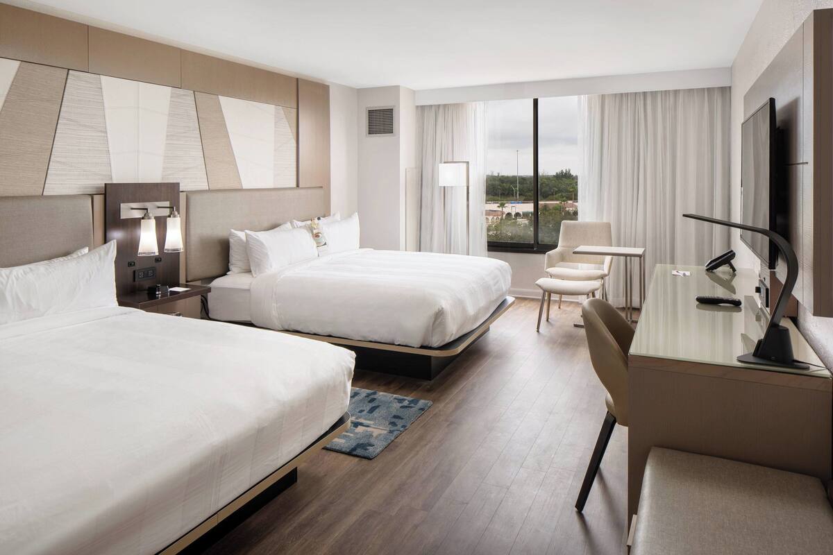 Een hotelkamer met twee bedden, een bureau, een stoel, een televisie en een groot raam met uitzicht.