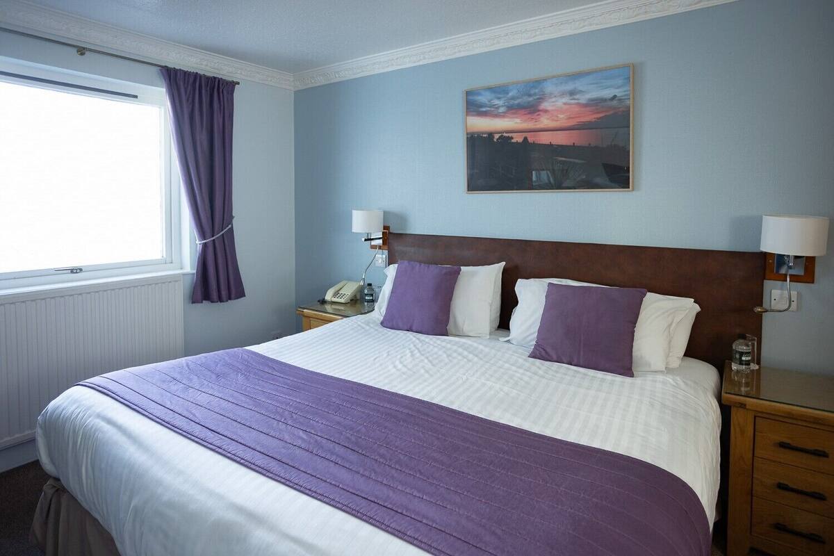 A hotel room with a large bed, two bedside tables, a window with curtains, a painting on the wall, and a nightstand with a lamp.