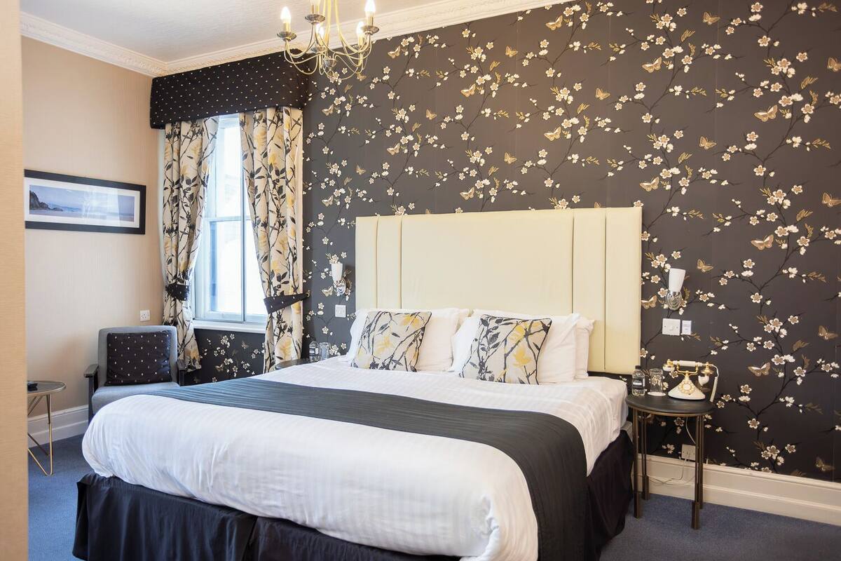 A hotel room with a large bed, floral wallpaper, a chandelier, and a vintage telephone on a side table.