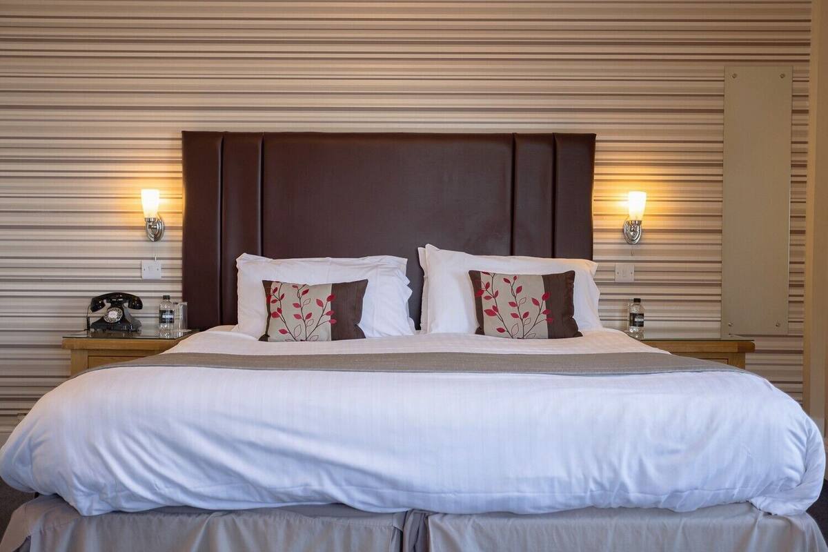 A neatly made bed with white linens and two decorative pillows, a wooden nightstand with a telephone and a bottle, and wall-mounted lamps on either side of the bed.