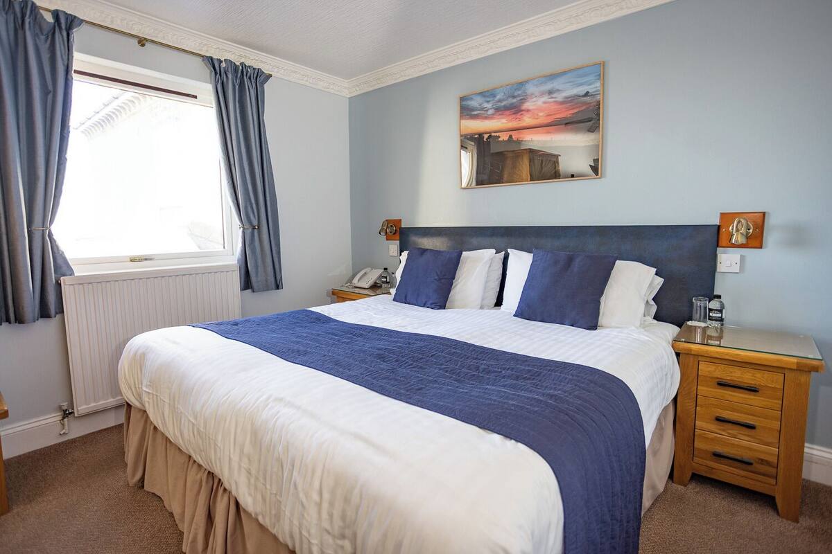 A neatly made bed with a blue and white duvet, a wooden nightstand with a lamp, and a painting of a sunset on the wall.