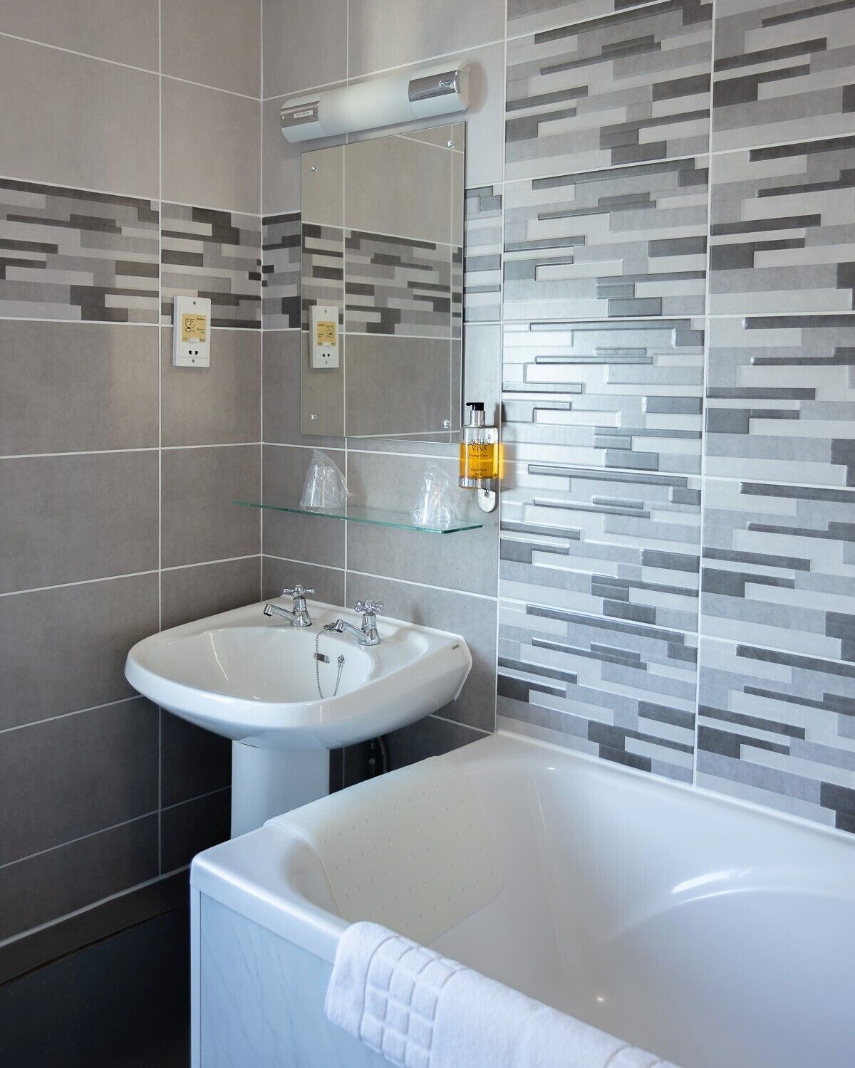 Ein modernes Badezimmer mit einem weißen Waschbecken, einer Badewanne und einem Spiegel. Zwei Steckdosen sind an der Wand angebracht, und eine Lotionflasche ist sichtbar.