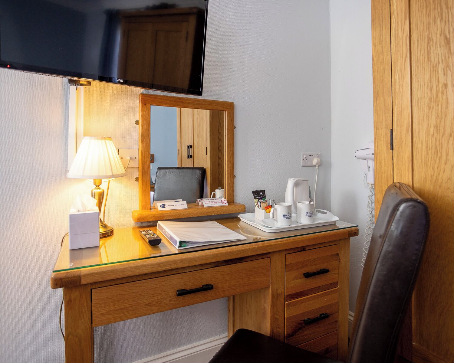 A hotel room with a wooden desk, a mirror, a lamp, a tray with coffee cups, a kettle, and a telephone.