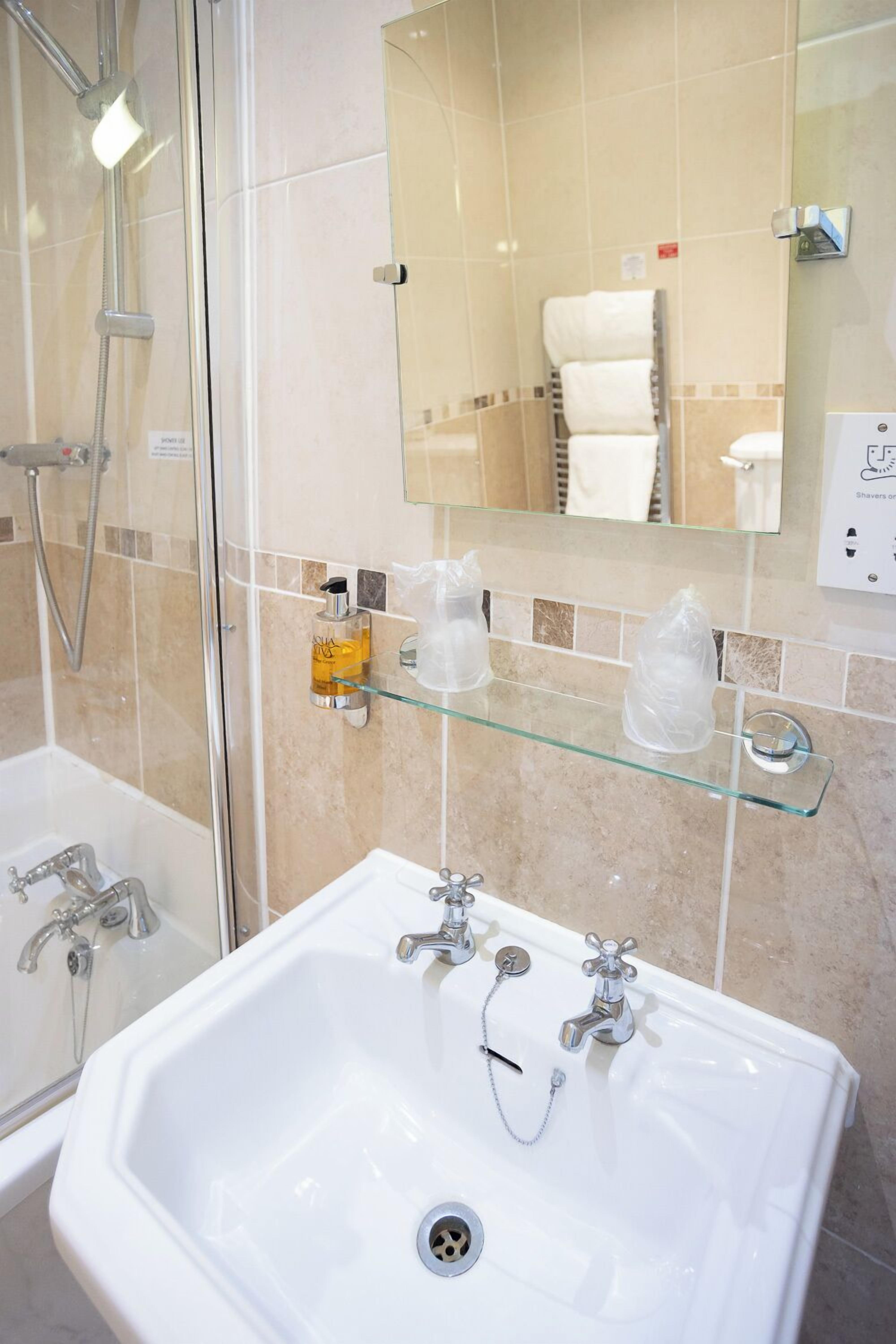 A bathroom with a glass shower enclosure, a white sink with two faucets, and a glass shelf with toiletries.