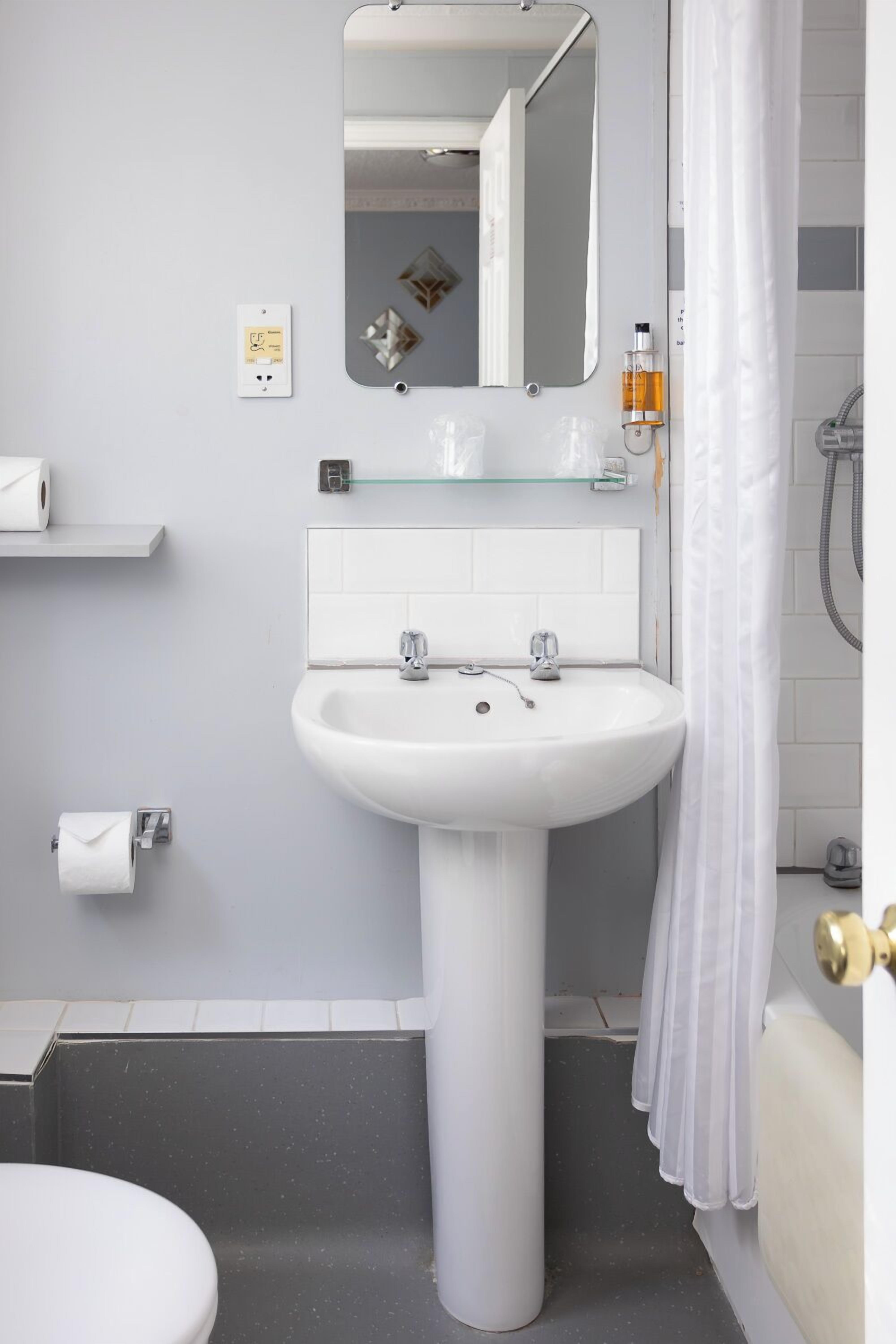 A bathroom with a pedestal sink, a mirror, a glass shelf with toiletries, a toilet, and a shower curtain.