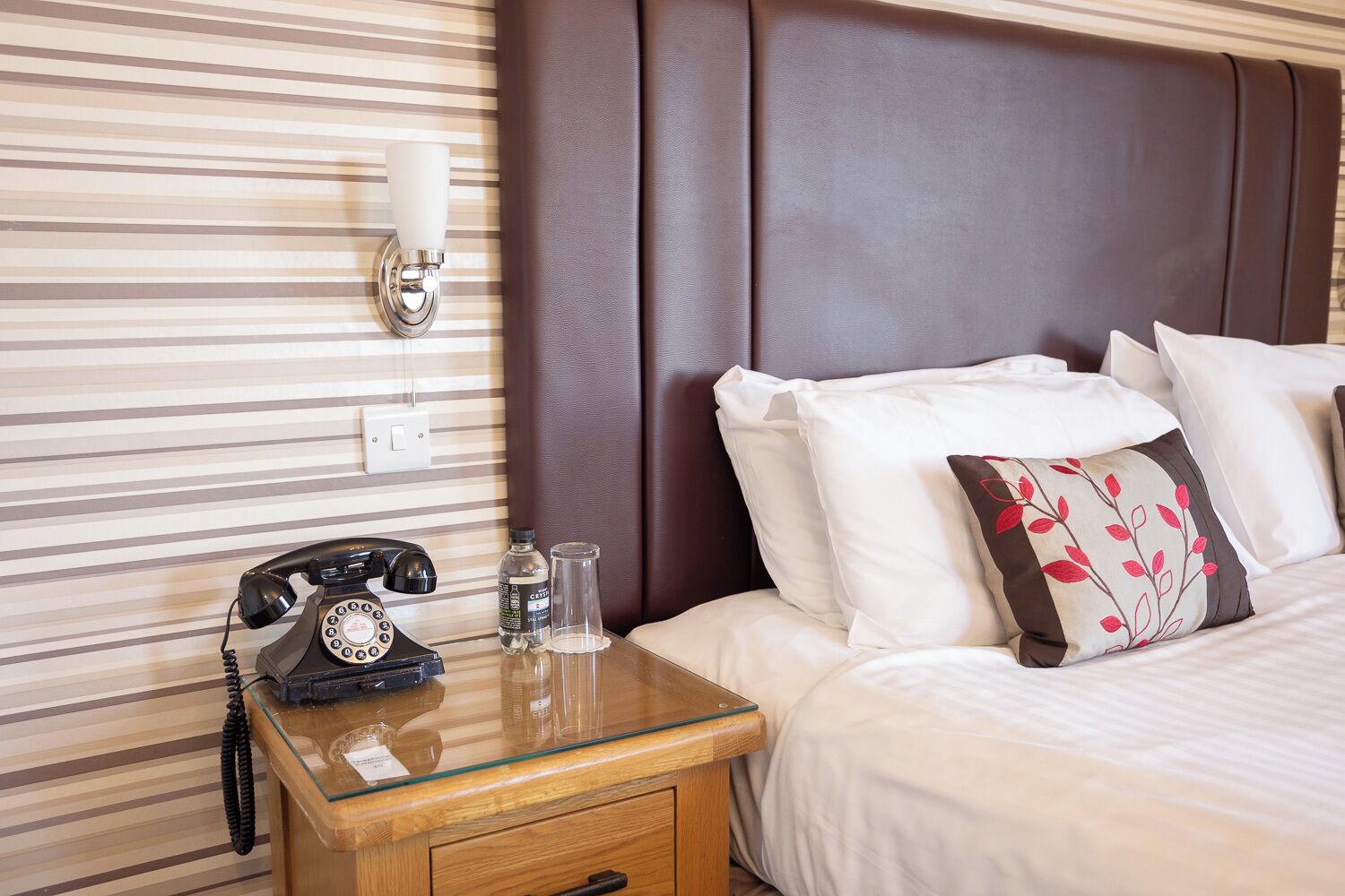 A hotel room with a striped wall, a wooden nightstand with a rotary phone, a glass table, a bed with white and patterned pillows, and a brown headboard.
