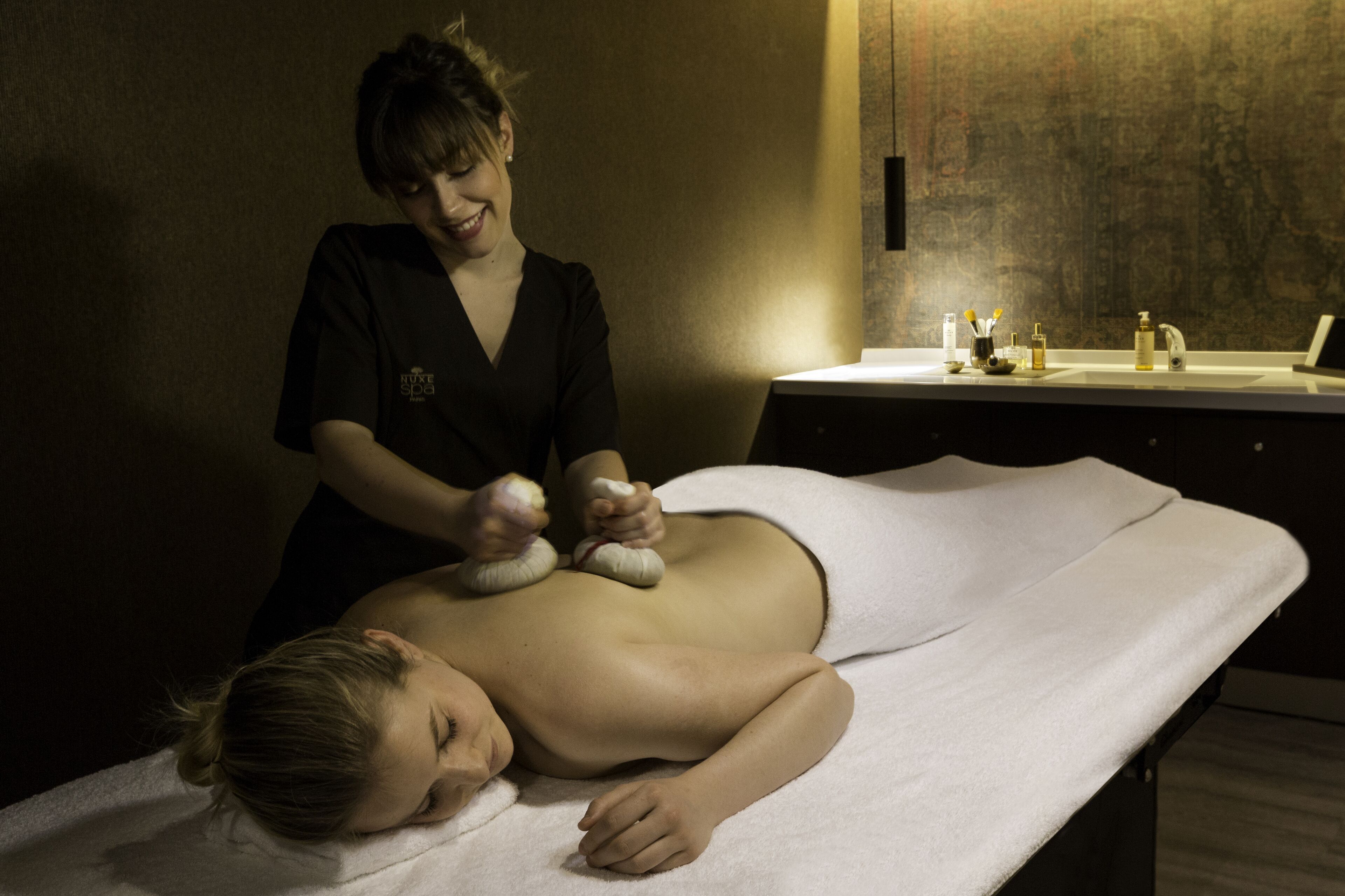 A massage therapy session in a dimly lit room with a massage table, a towel, and a sink with toiletries.